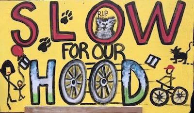 A hand painted sign reading "Slow for our hood" depicting children, pets, wldlife, and a cyclist.